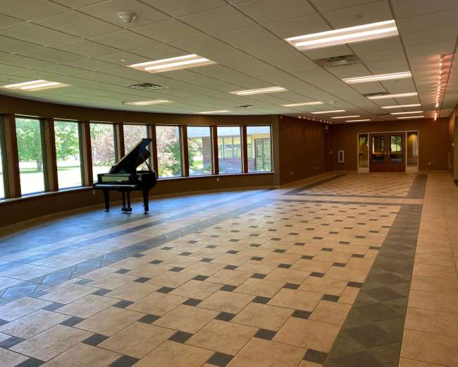 An empty room with a grand piano on one side and large windows lining the walls, allowing natural light to illuminate the tile floor.