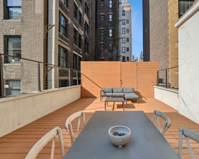 The image shows a rooftop terrace with wooden flooring, a table with a bowl, and a gray couch, surrounded by tall buildings.