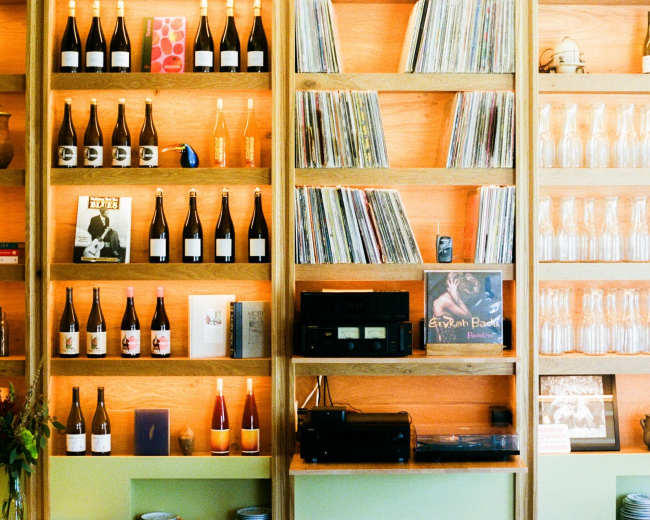 A wooden shelf displays bottles of wine, vinyl records, and glass containers against an orange wall.