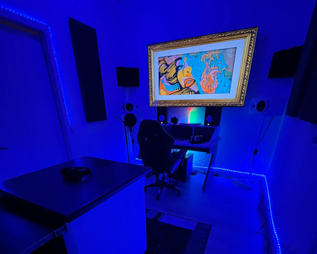 The image shows a dimly lit room with blue LED lights, featuring a desk with audio equipment and a framed artwork on the wall.