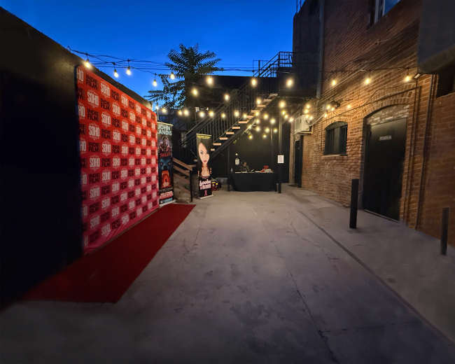 The image shows a dimly lit alleyway decorated with string lights, featuring a red carpet leading to a promotional backdrop and a table in the background.