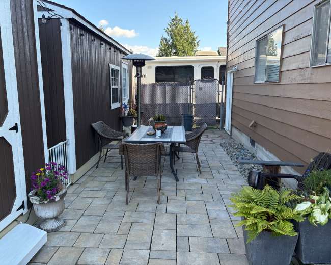 The image shows a backyard patio area with a table and four chairs, surrounded by decorative plants and a storage shed, with a trailer partially visible in the background.