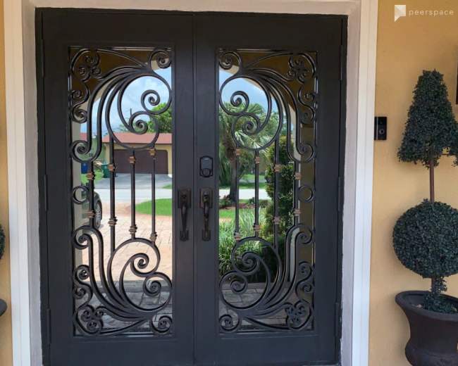 A pair of ornate double doors with intricate ironwork designs set against a yellow wall, flanked by potted topiary plants.