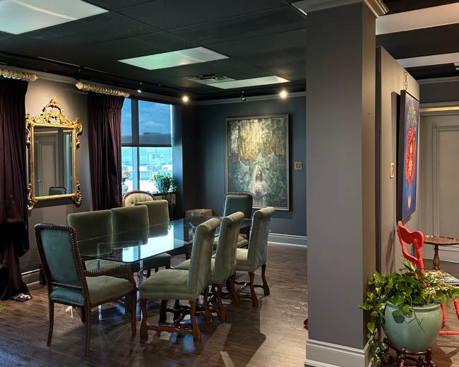 The image shows a modern, well-furnished meeting room featuring a glass table surrounded by green upholstered chairs, with dark walls and large windows letting in natural light.