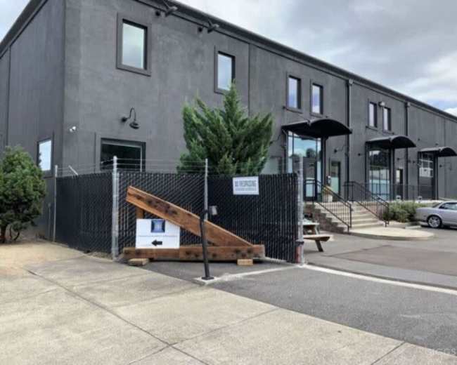 The image shows a gray building with a fenced entrance featuring a wooden ramp and a sign directing visitors.