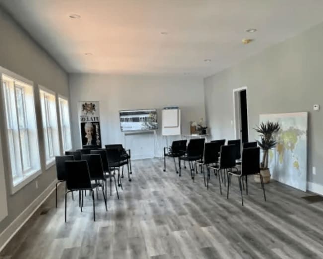 The image shows a brightly lit, minimalist room with several black chairs arranged in rows and a large artwork on the wall.