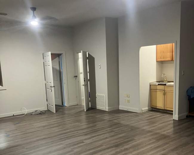 The image shows a spacious room with wooden flooring, two open doors leading to separate areas, and a kitchenette visible in an alcove.