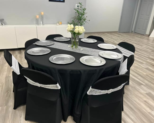 A round table is set for a formal meal with silver plates and a black tablecloth, surrounded by eight chairs draped in black covers with silver accents.