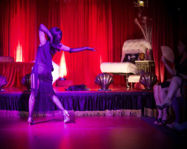A dancer performs on stage in front of a red curtain, with a chaise lounge and audience seated nearby.