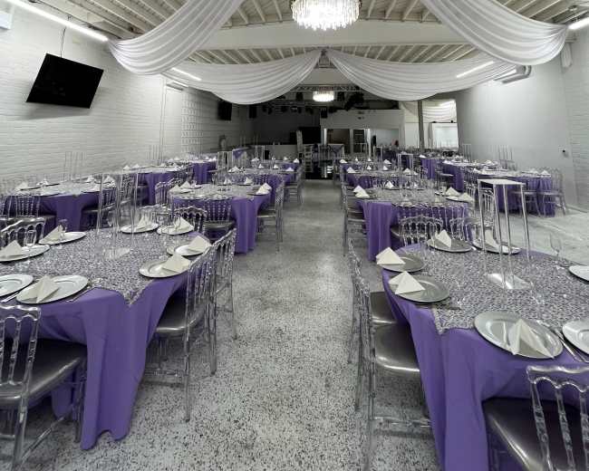 The image shows a large event space set up for a formal gathering, featuring several tables covered with purple tablecloths and adorned with elegant place settings under a draped ceiling and a chandelier.