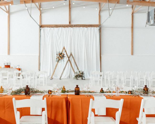 A long table with orange linens is set up in a spacious event venue, featuring a backdrop of white drapes and a wooden frame adorned with greenery and flowers.