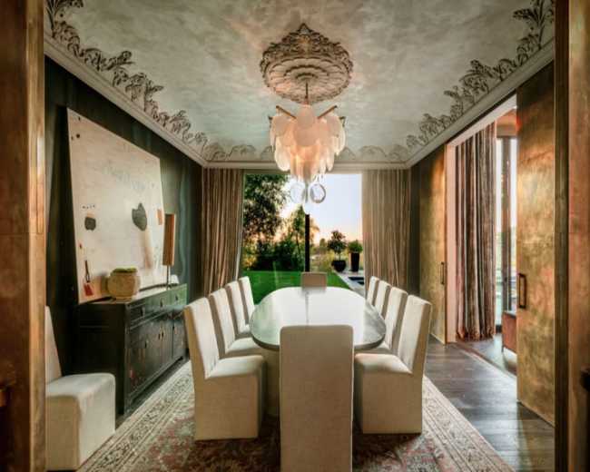 The image shows an elegant dining room featuring a large oval table surrounded by upholstered chairs, with a decorative ceiling and a chandelier above, and large glass doors leading to a garden view.
