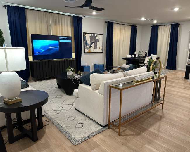 The image shows a modern living room featuring a light-colored sofa, a black coffee table, a large television, and blue curtains.