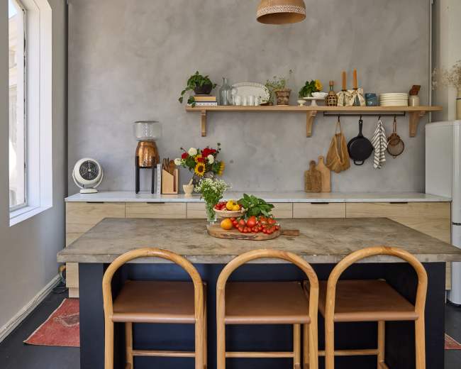The kitchen features a wooden island with three chairs, a marble countertop, and an arrangement of fresh produce and flowers, alongside open shelving and a vintage refrigerator.