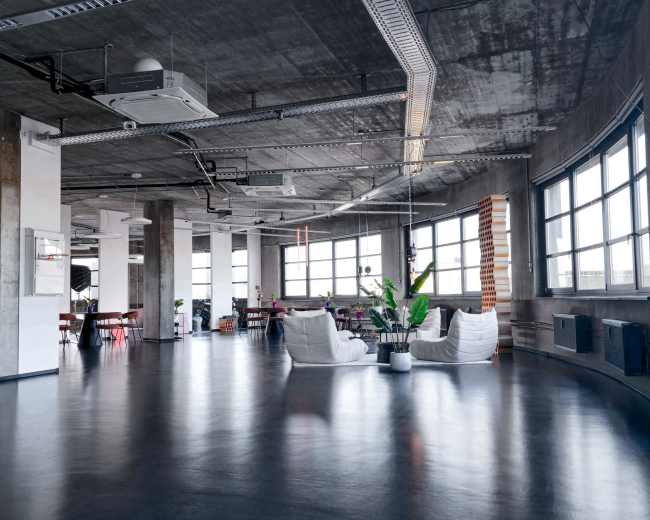The image shows a modern, open office space with large windows, concrete walls, and minimalist furniture.