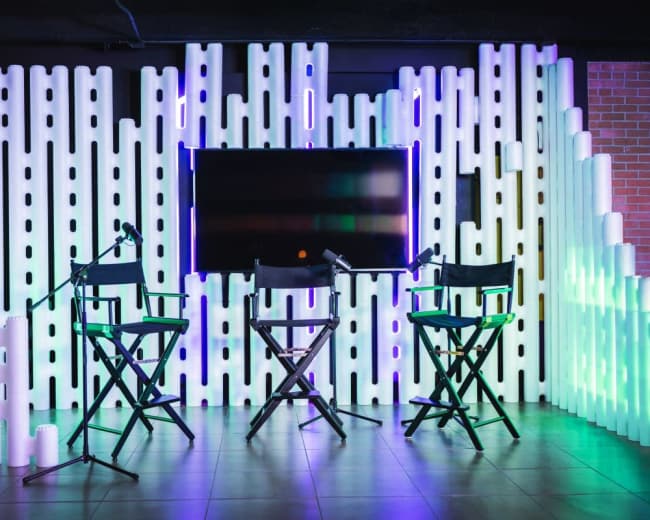 The scene features three director's chairs arranged in front of a television on a backdrop made of vertical white tubes illuminated with colorful lights.