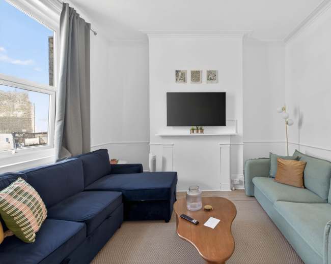 The image shows a modern living room with two sofas, a wooden coffee table, a TV mounted on the wall, and large windows.
