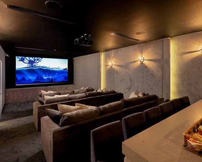 The image shows a home theater room with several rows of plush seating facing a large screen, illuminated by wall sconces and a snack table in the foreground.