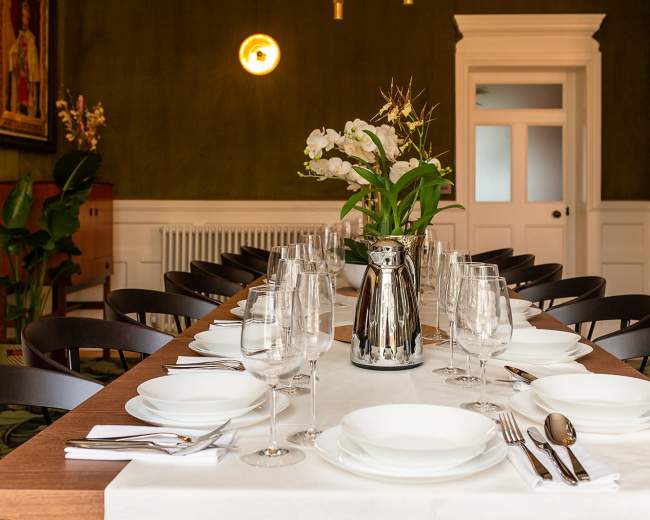 A large dining table is set with white plates, glassware, and silverware, accompanied by a floral arrangement in a modern vase, all situated in a room with green walls and elegant decor.