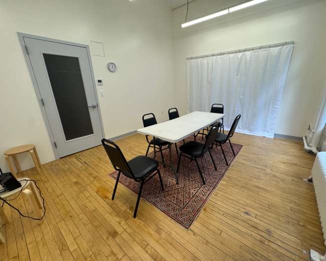 The image shows a simple meeting room with a rectangular table surrounded by six chairs, a stool in the corner, a wall clock, and a curtain covering a window.