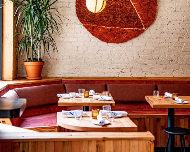 The image shows an interior of a modern restaurant featuring a circular red wall mural, wooden tables, and a cozy banquette seating area with plants.