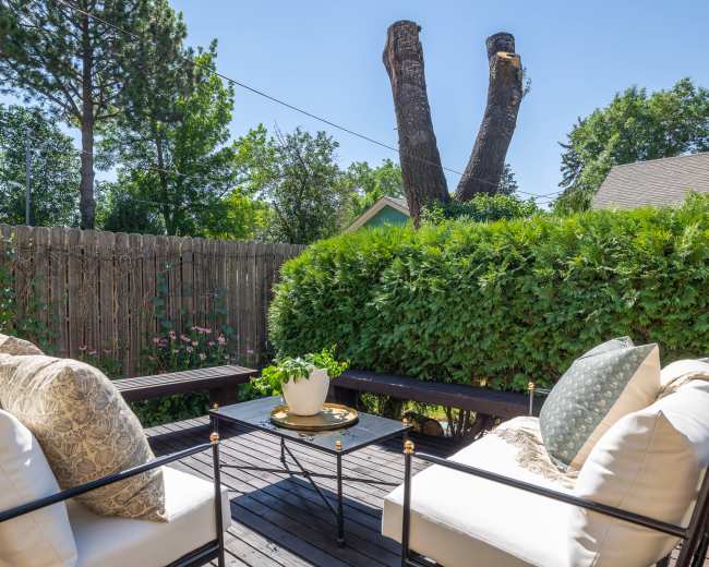 A seating area with white cushions and a small table is set on a wooden deck surrounded by greenery and a wooden fence, with two tall, cut trees in the background.
