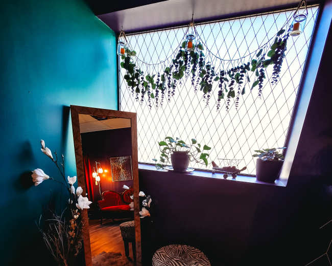 A cozy corner features a large mirror, a patterned stool, a faux fur rug, and potted plants near a window draped with greenery.