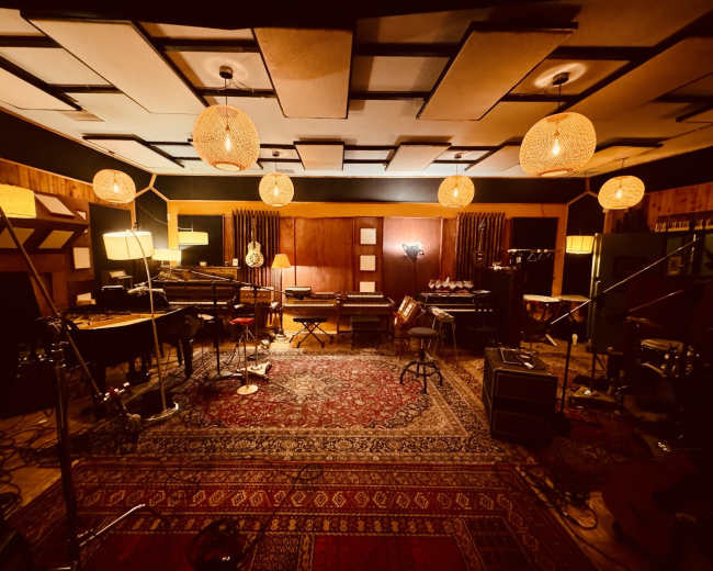 The image shows a well-lit music recording studio featuring an array of musical instruments, sound equipment, and various lighting fixtures.