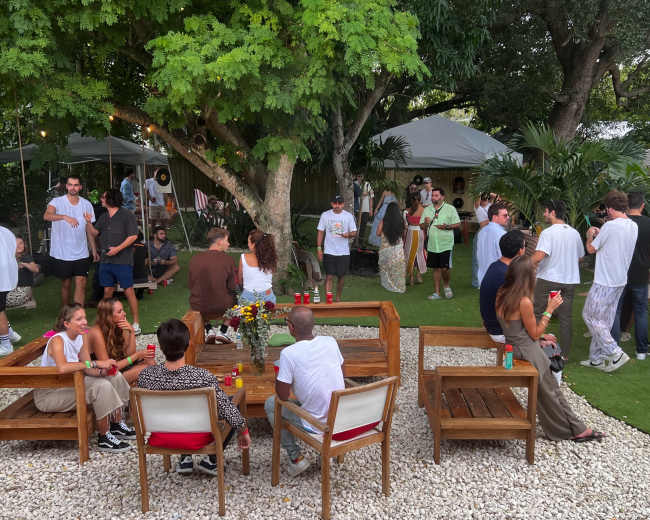 A garden party scene features groups of people socializing around wooden furniture, surrounded by lush greenery and decorative elements.