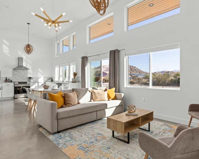 A modern living room features a light-colored sofa with yellow accent pillows, a wooden coffee table, and large windows providing a view of the outdoors.