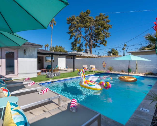 A backyard swimming pool features colorful floats, lounge chairs, and umbrellas, surrounded by a landscaped area with palm trees and a modern home.