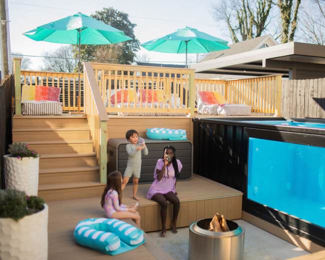 A family enjoys a sunlit backyard with a wooden deck, pool, and seating areas beneath green umbrellas.