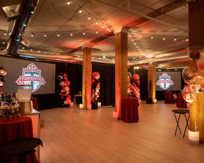 The image shows a decorated event space with tables draped in red fabric, string lights overhead, and large screens displaying a logo, surrounded by festive balloon arrangements.