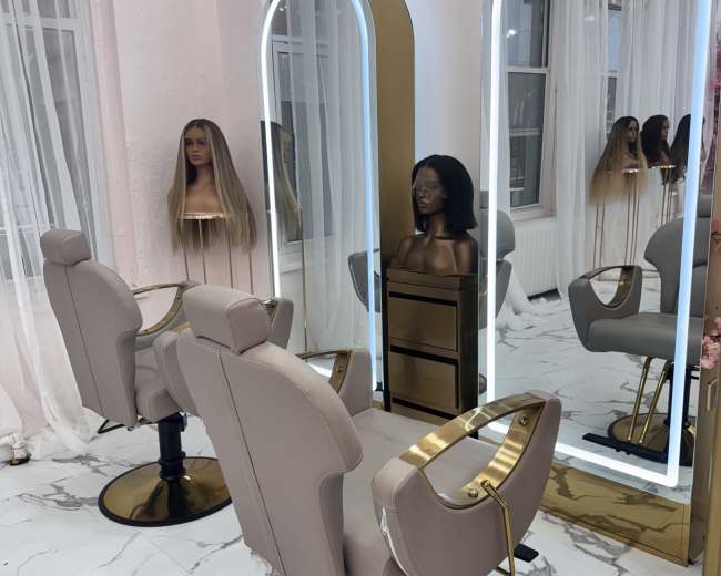 The image shows a modern hair salon interior featuring two stylish chairs, illuminated mirrors, and mannequin heads with wigs displayed on the wall.