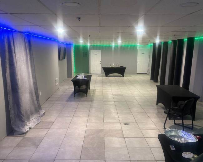 The image shows a spacious event room with gray walls, tiled flooring, and several black tables arranged throughout, illuminated by green and blue LED lights along the ceiling.