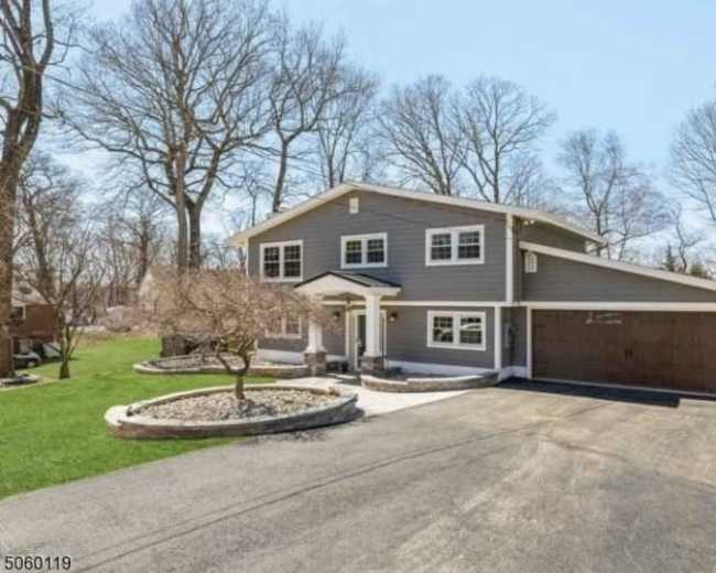 The image shows a two-story gray house with a garage, situated on a landscaped property with trees and a clear blue sky.