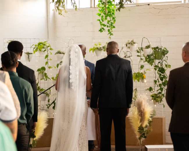 A couple stands at the altar surrounded by greenery, while guests look on during a wedding ceremony.