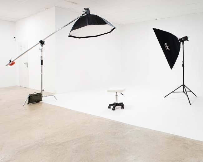 A minimalist photography studio features a white backdrop, a stool, and lighting equipment set up on a concrete floor.