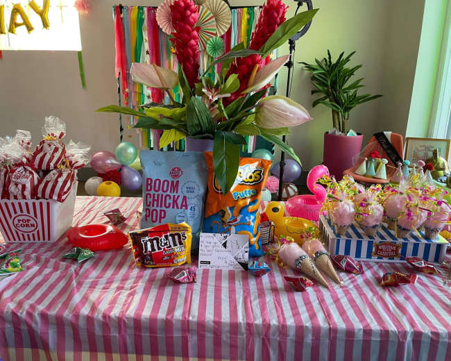 A colorful party table is decorated with snacks and treats, including popcorn, candies, and floral arrangements.