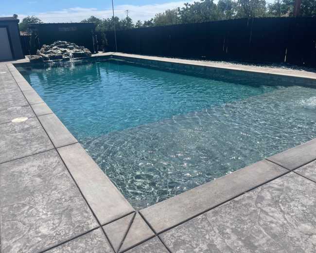 The image shows a rectangular swimming pool with a textured concrete deck, surrounded by a black wooden fence and greenery in the background.
