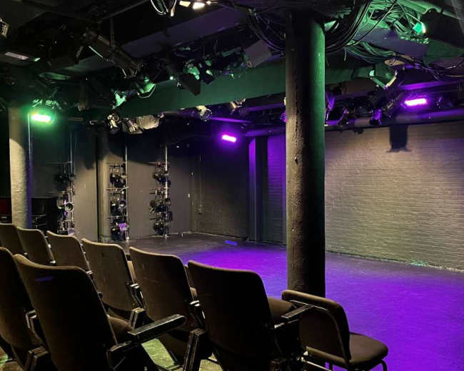 The image shows an empty theater stage with rows of black chairs facing a well-lit performance area featuring colorful stage lights.