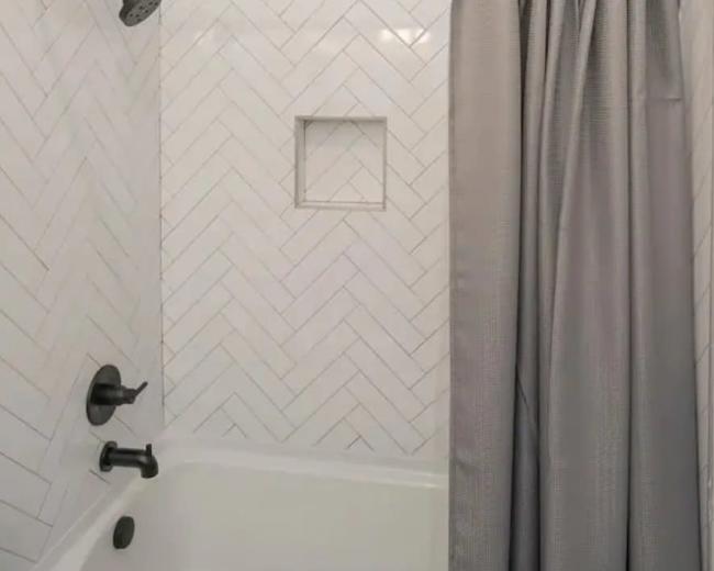 The image shows a bathroom with a white tiled shower featuring a herringbone pattern, a curved shower rod with a gray curtain, and a built-in shelf for toiletries.