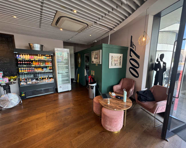 The image shows a coffee shop interior with a beverage display fridge, a small seating area featuring two pink chairs, and large glass doors leading outside.