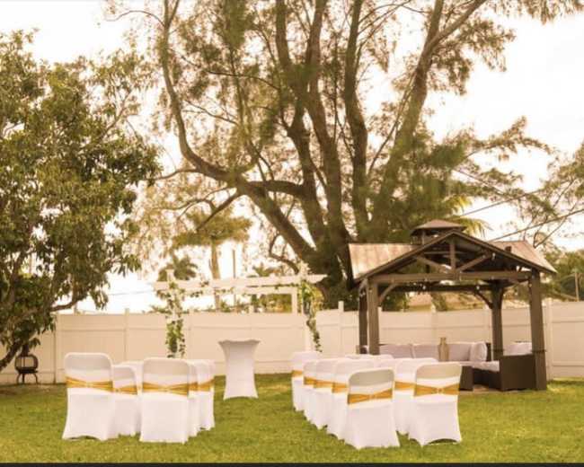 The image shows a wedding setup in a green outdoor area, featuring rows of white chairs with gold sashes, a gazebo, and a small table.