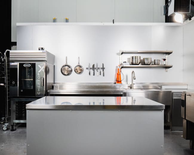 The image shows a modern kitchen setup featuring stainless steel counters, a baking oven on the left, and various kitchen utensils hanging on the wall.