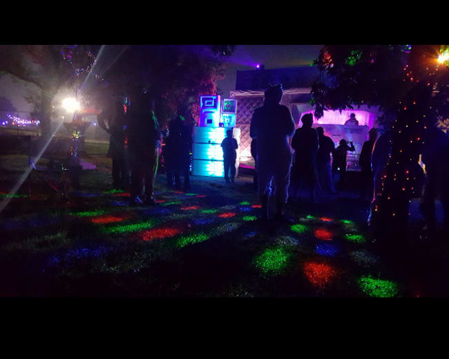 A crowd of people gathers in a dimly lit outdoor area with colorful lights illuminating the ground.
