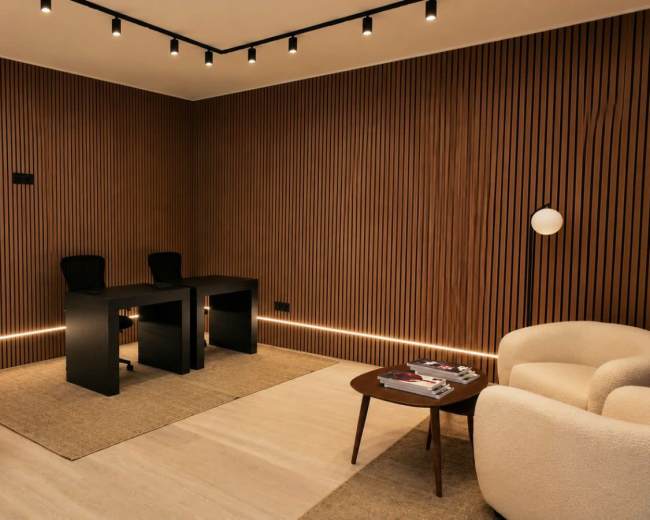 The image shows a minimalist office space with two black desks, matching chairs, and a round coffee table, all set against wooden paneled walls and soft lighting.