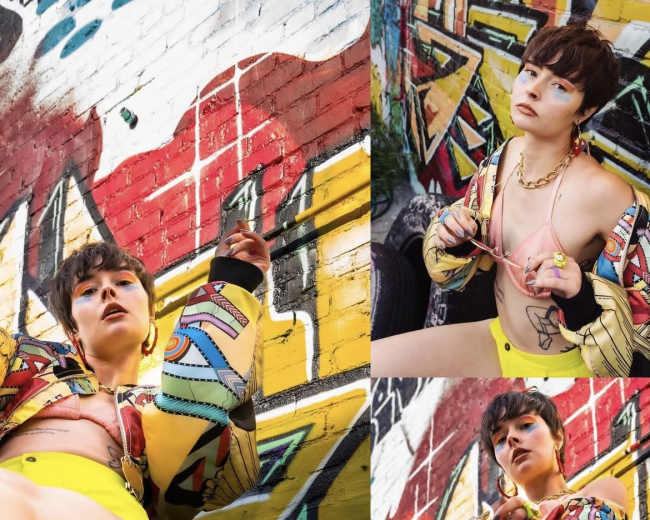 A person wearing a colorful jacket and shorts poses on a barrel in front of a vividly painted graffiti wall.