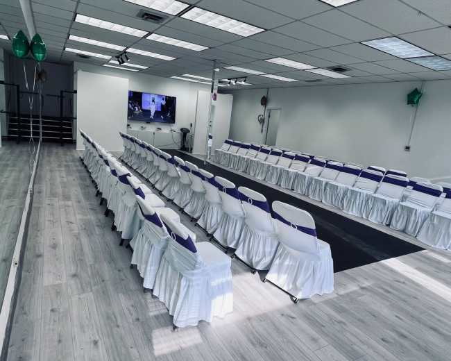 A spacious room features rows of white chairs with purple accents arranged for an event, along with a mirrored wall and modern audiovisual equipment.