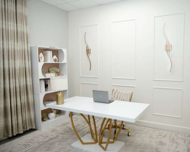 A modern office space features a white desk with a laptop, a decorative shelf, and subtle wall panels in a neutral-toned room.
